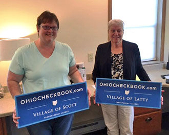 Latta Fiscal Officer Kay Miller and Scott Fiscal Officer Hilary Yoder with signs noting their villages' participation in the Ohio Checkbook.com project. (photo submitted)